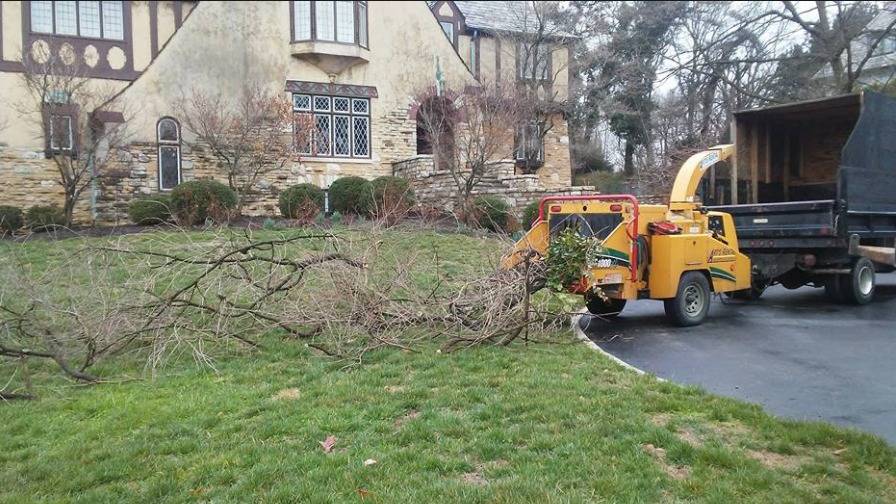 A yellow wood chipper and a dump truck at the edge of a driveway beside a stone Tudor-style house, with cut branches piled on the lawn.